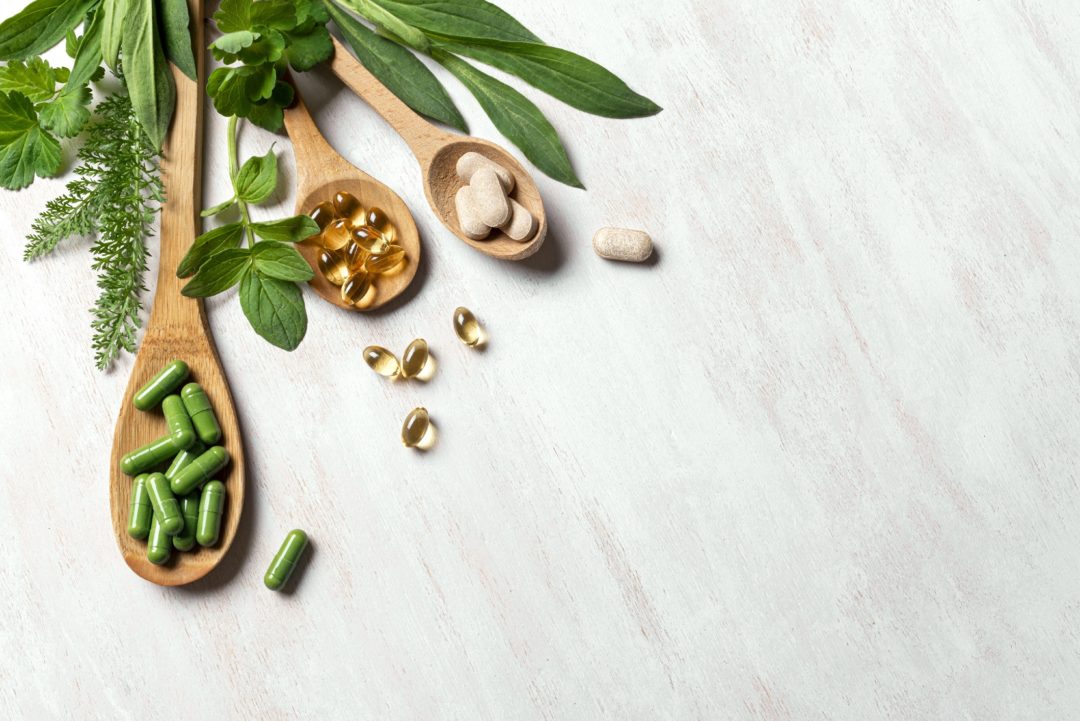 Natural vitamins, plant based supplements and green leaves on white wooden background, top view.