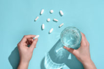 Woman holding in hands white pill and glass of water on light blue background. Top view, fat lay. 