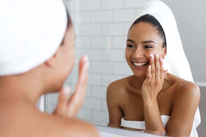 Happy woman putting on facial moisturizer with hand in bathroom mirror