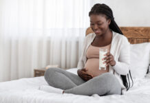 Healthy Calcium Drink. Happy Black Pregnant Woman Holding Glass Of Milk, Sitting On Bed At Home And Touching Her Belly, Expectant African Woman Restng In Bedroom Enjoying Pregnancy Time, Copy Space