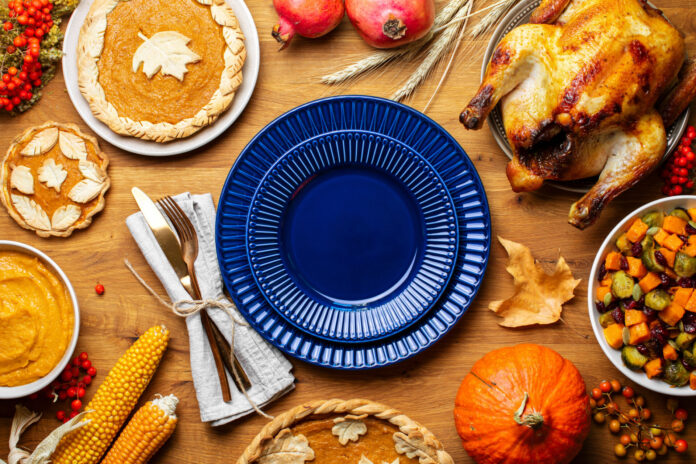 A blue plate on a brown wood table. A fork and knife are on a napkin next to the plate. The plate is surrounded by traditional thanksgiving foods and general fall concepts, including a cooked turkey, several pies, a pumpkin, a bowl of chopped squash vegetables, and a dead leaf.