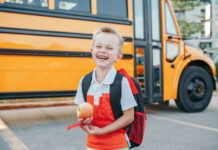 Happy laughing smiling Caucasian boy student by yellow bus on first September day. Hard of hearing child kid eating apple fruit at school yard outdoor. Education and back to school in Autumn Fall.