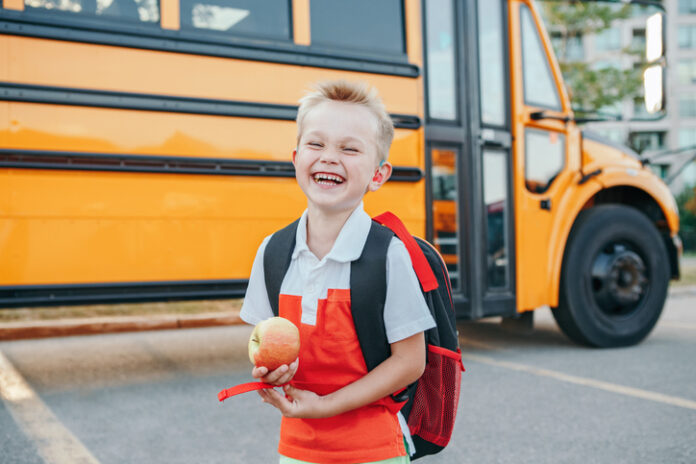 Happy laughing smiling Caucasian boy student by yellow bus on first September day. Hard of hearing child kid eating apple fruit at school yard outdoor. Education and back to school in Autumn Fall.