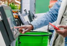 supermarket checkout selects the desired product on the electronic screen of the cash register with a phone in his hands against the background of green shopping baskets, retail and self-service checkout in a hypermarket