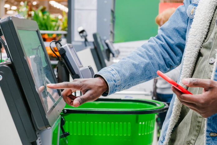 supermarket checkout selects the desired product on the electronic screen of the cash register with a phone in his hands against the background of green shopping baskets, retail and self-service checkout in a hypermarket
